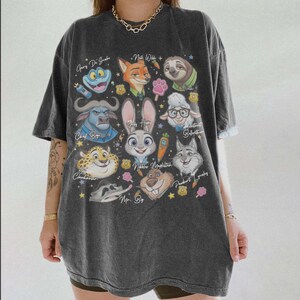 K&ouml;nnte beinhalten: Ein anthrazitfarbenes Kurzarm-T-Shirt mit einem Grafikdruck verschiedener animierter Tierfiguren aus Zootopia, darunter Judy Hopps, Nick Wilde und Flash, deren Namen in Schreibschrift neben kleinen Sternen und Symbolen aufgedruckt sind.
