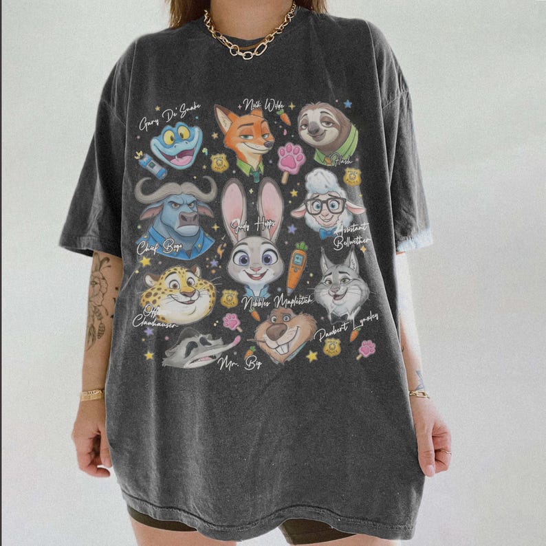 K&ouml;nnte beinhalten: Ein anthrazitfarbenes Kurzarm-T-Shirt mit einem Grafikdruck verschiedener animierter Tierfiguren aus Zootopia, darunter Judy Hopps, Nick Wilde und Flash, deren Namen in Schreibschrift neben kleinen Sternen und Symbolen aufgedruckt sind.