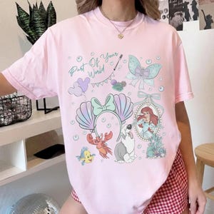 May include: A light pink short-sleeve t-shirt featuring a graphic print with the text Part Of Your World, a mermaid, a crab, a fish, a dog, a seashell headband, a butterfly, and bubbles in shades of blue, purple, red, and white.