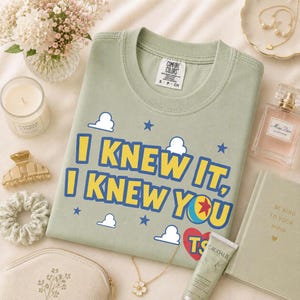 Pu&ograve; includere: Una t-shirt girocollo color verde salvia con la scritta gialla I KNEW IT, I KNEW YOU con contorni blu, una grafica a palla colorata, icone di nuvole bianche, stelle blu e un cuore rosso con le lettere TS stampate sul petto.