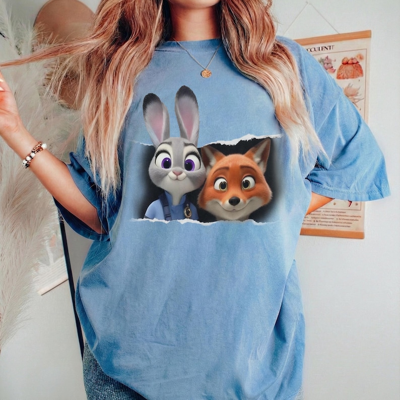 K&ouml;nnte beinhalten: Hellblaues T-Shirt mit einem Aufdruck von Judy Hopps und Nick Wilde aus Zoomania. Judy ist ein graues Kaninchen mit lila Augen, und Nick ist ein oranger Fuchs. Der Aufdruck scheint durch einen Riss im T-Shirt zu blicken.