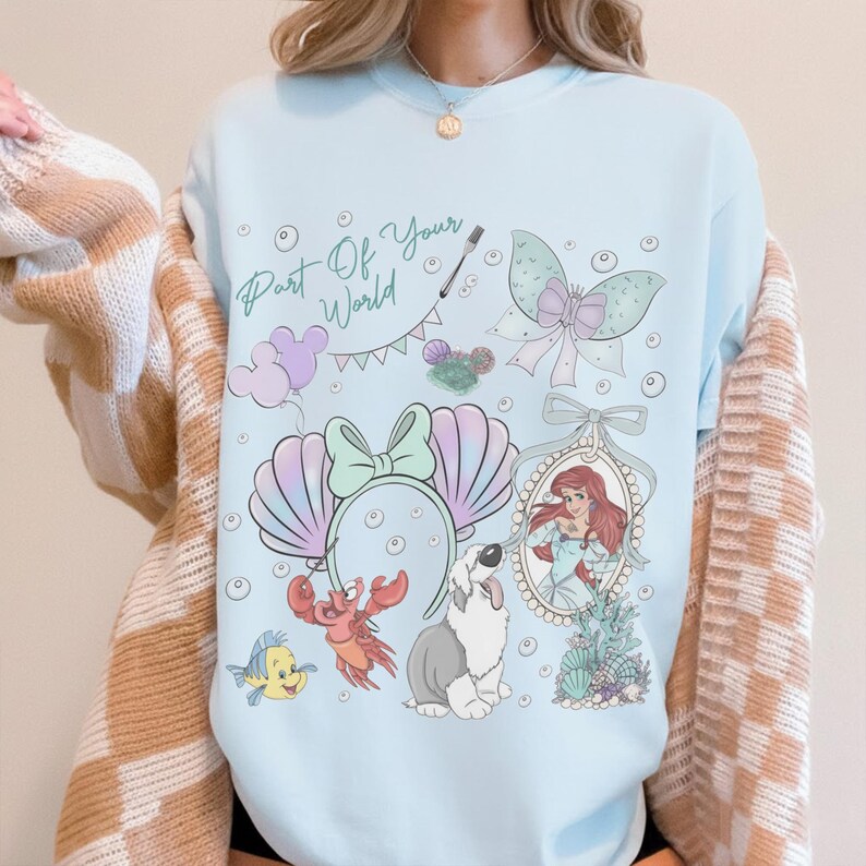 May include: A light blue crewneck sweatshirt featuring a graphic print with the text Part Of Your World, a mermaid, a crab, a fish, a dog, seashell ears, a bow, and bubbles. The design includes pastel colors and various underwater themed icons.