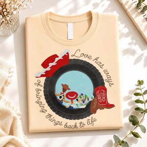 May include: A light beige t-shirt featuring a graphic of a black tire framing a scene with three cartoon characters. A red cowboy hat sits on the tire, and a red cowboy boot with the name Taylor is beside it. The text reads Love has ways of bringing things back to life.
