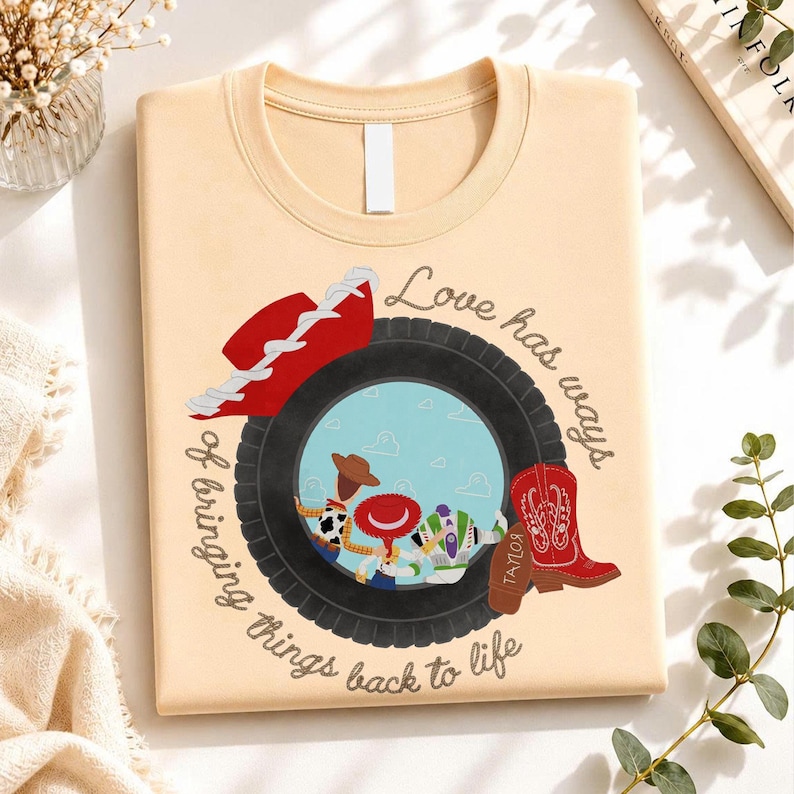 May include: A light beige t-shirt featuring a graphic of a black tire framing a scene with three cartoon characters. A red cowboy hat sits on the tire, and a red cowboy boot with the name Taylor is beside it. The text reads Love has ways of bringing things back to life.