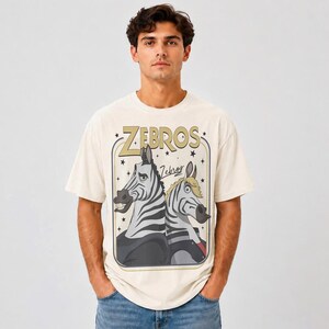 K&ouml;nnte beinhalten: Ein cremefarbenes Kurzarm-T-Shirt mit einem Grafikdruck von zwei Zeichentrick-Zebras. Der Text "ZEBROS" erscheint in gro&szlig;en goldenen Buchstaben &uuml;ber den Zebras, mit dem Wort "Zebros" in Schreibschrift darunter. Das Design ist in einem Rahmen.