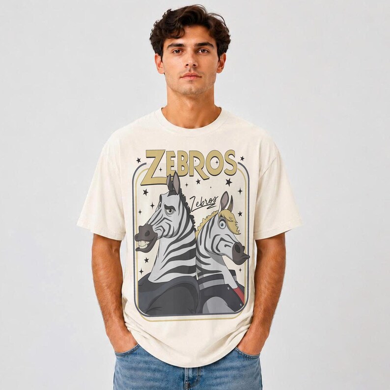 K&ouml;nnte beinhalten: Ein cremefarbenes Kurzarm-T-Shirt mit einem Grafikdruck von zwei Zeichentrick-Zebras. Der Text "ZEBROS" erscheint in gro&szlig;en goldenen Buchstaben &uuml;ber den Zebras, mit dem Wort "Zebros" in Schreibschrift darunter. Das Design ist in einem Rahmen.