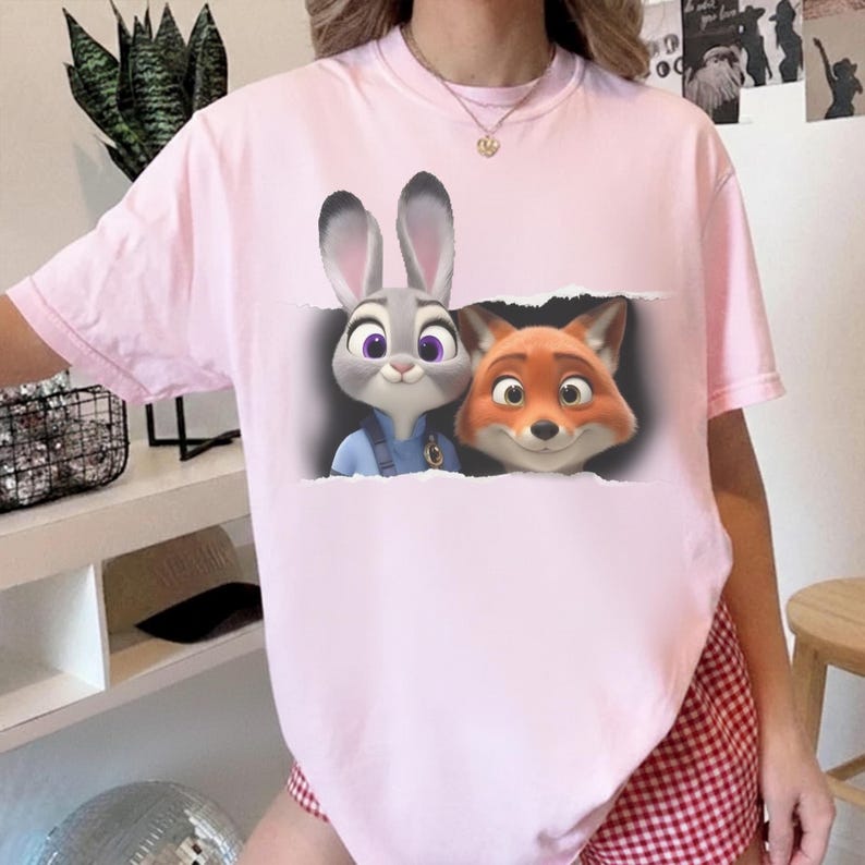 K&ouml;nnte beinhalten: Hellrosa T-Shirt mit einem Aufdruck von Judy Hopps und Nick Wilde aus Zoomania. Judy, ein graues Kaninchen, und Nick, ein oranger Fuchs, l&auml;cheln. Das Shirt ist ein l&auml;ssiger Stil mit kurzen &Auml;rmeln.