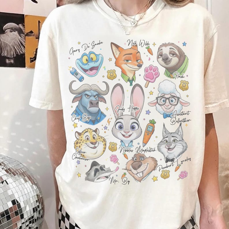 K&ouml;nnte beinhalten: Ein cremefarbenes T-Shirt mit einem Grafikdruck verschiedener animierter Charaktere aus Zoomania, darunter Judy Hopps, Nick Wilde, Flash, Chief Bogo und andere, begleitet von ihren Namen und kleinen Symbolen wie Karotten und Polizeimarken.