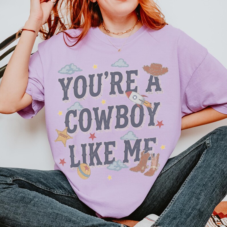 May include: A light purple t-shirt featuring the dark blue text "YOU'RE A COWBOY LIKE ME" surrounded by graphic illustrations of a brown cowboy hat, a rocket ship, a yellow sheriff star, a ball, brown cowboy boots, and light blue clouds.