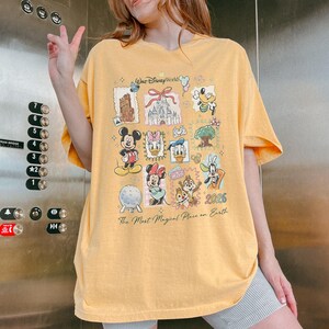 May include: Yellow t-shirt featuring a collage of Disney characters and landmarks, including Mickey Mouse, Minnie Mouse, and Cinderella's castle. The shirt has the text "Walt Disney World" and "2026" printed on it. The phrase "The Most Magical Place on Earth" is also printed on the shirt.