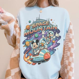 Może przedstawiać: Jasnoniebieska bluza z nadrukiem w stylu retro przedstawiającym Myszka Miki, Kaczora Donalda i Goofiego w skafandrach kosmicznych na rakiecie. Napis "SPACE MOUNTAIN" znajduje się nad postaciami. Projekt zawiera planety, gwiazdy i kosmiczne tło.