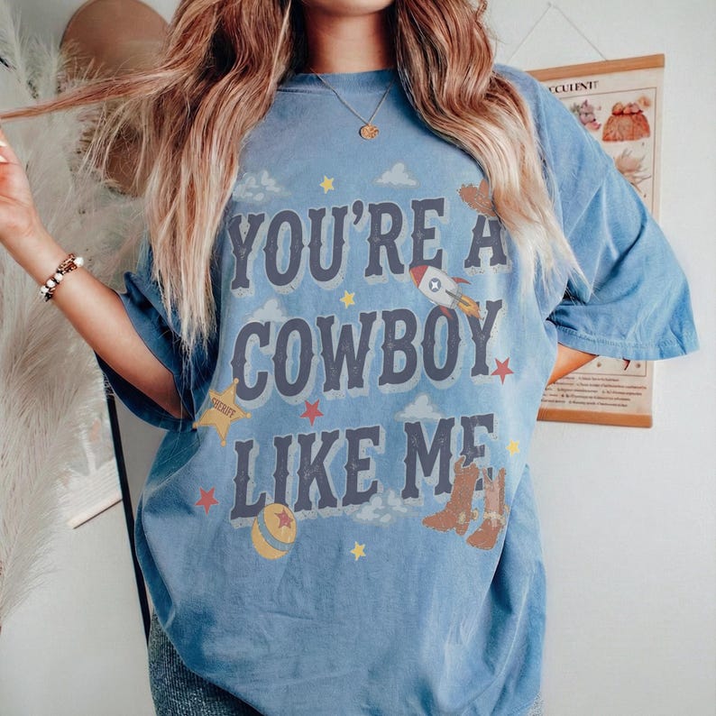 May include: A blue short-sleeve t-shirt featuring the text "YOU'RE A COWBOY LIKE ME" in dark blue letters. The design includes graphic illustrations of a rocket, a sheriff star, a ball, and cowboy boots, accented with small stars and clouds.