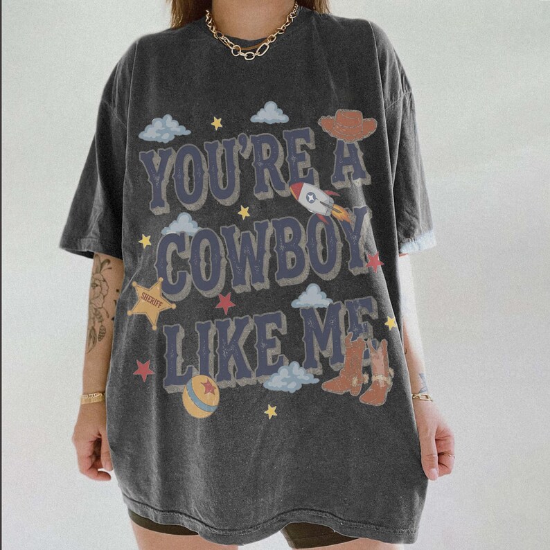 May include: A charcoal gray oversized t-shirt featuring the text "YOU'RE A COWBOY LIKE ME" in a distressed blue font. The graphic includes illustrations of a rocket, a sheriff star, cowboy boots, a hat, a ball, and small clouds and stars.