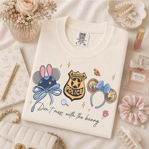 May include: A cream-colored crewneck t-shirt featuring a graphic print of Zootropolis-themed mouse ears, a police badge, and a carrot. The text "Don't mess with the bunny" is printed in black script below the illustrations on the front of the shirt.
