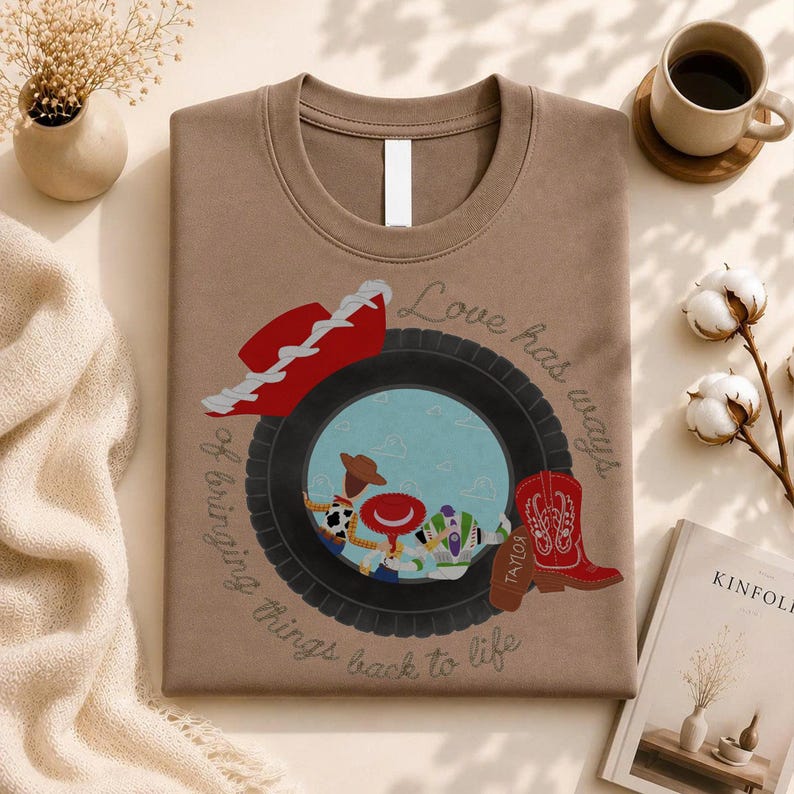 May include: A brown t-shirt featuring a graphic of a black tire framing a scene with cartoon characters, a red cowboy hat, and a red cowboy boot. The text "Love has ways of bringing things back to life" is printed in a cursive font around the tire.