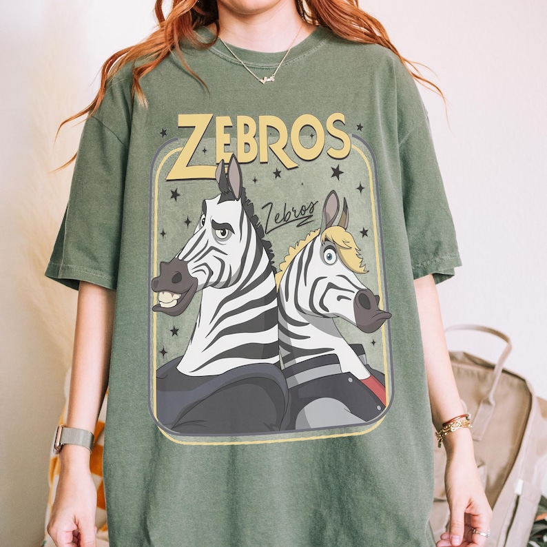 K&ouml;nnte beinhalten: Ein olivgr&uuml;nes Kurzarm-T-Shirt mit einem Grafikdruck von zwei Zeichentrick-Zebras. Der Text "ZEBROS" erscheint in gro&szlig;en gelben Buchstaben &uuml;ber den Zebras, mit einem kleineren "Zebros"-Schriftzug darunter. Das Design enth&auml;lt Sterne und einen Rahmen.