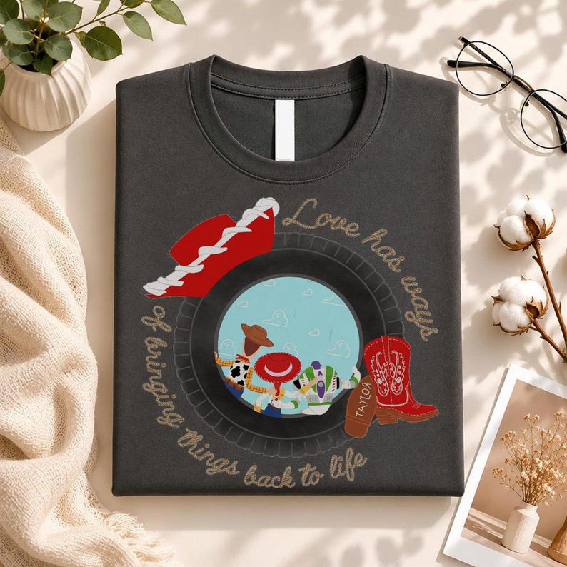 May include: A dark gray t-shirt featuring a circular graphic with characters from Toy Story, a red cowboy hat, and a red cowboy boot. The text "Love has ways of bringing things back to life" is printed in a cursive font around the central circular design.