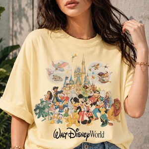 May include: A light yellow t-shirt featuring a colorful graphic of various Disney characters, including Mickey Mouse, Donald Duck, and Simba, in front of a castle. The words "Walt Disney World" are printed in a script font at the bottom.