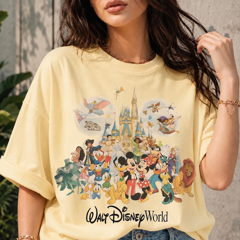 May include: A light yellow t-shirt featuring a colorful graphic of various Disney characters, including Mickey Mouse, Donald Duck, and Simba, in front of a castle. The words "Walt Disney World" are printed in a script font at the bottom.