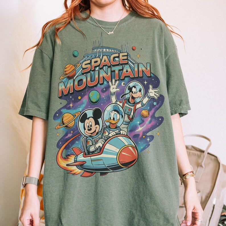 Może przedstawiać: Zielona koszulka z kolorową grafiką Myszki Miki, Kaczora Donalda i Goofy'ego w skafandrach kosmicznych na statku kosmicznym, z napisem "SPACE MOUNTAIN". Projekt zawiera planety i gwiezdne tło.