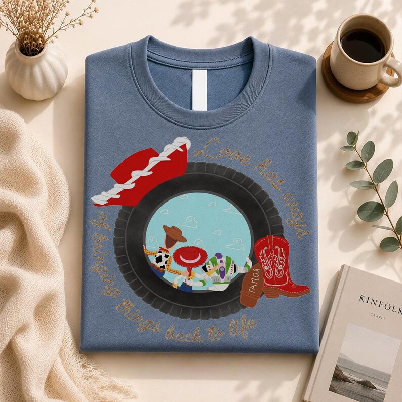 May include: A blue crewneck t-shirt featuring a graphic of a black tire framing a scene with three cartoon characters. A red cowboy hat sits above the tire, and a red cowboy boot is on the right. The text reads, "Love has ways of bringing tires back to life."