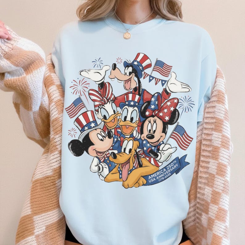 May include: Pale blue sweatshirt with a graphic of Disney characters Mickey, Minnie, Donald, Daisy, Goofy, and Pluto celebrating the 250th USA Anniversary. The design includes American flags and fireworks.