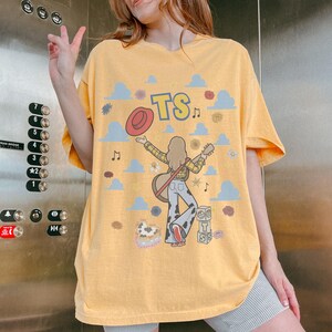 May include: A yellow t-shirt featuring a graphic print of a person with long hair holding a guitar, wearing a yellow plaid shirt and cow-print trousers. The design includes a red hat, blue clouds, musical notes, flowers, and blocks with the text TS.