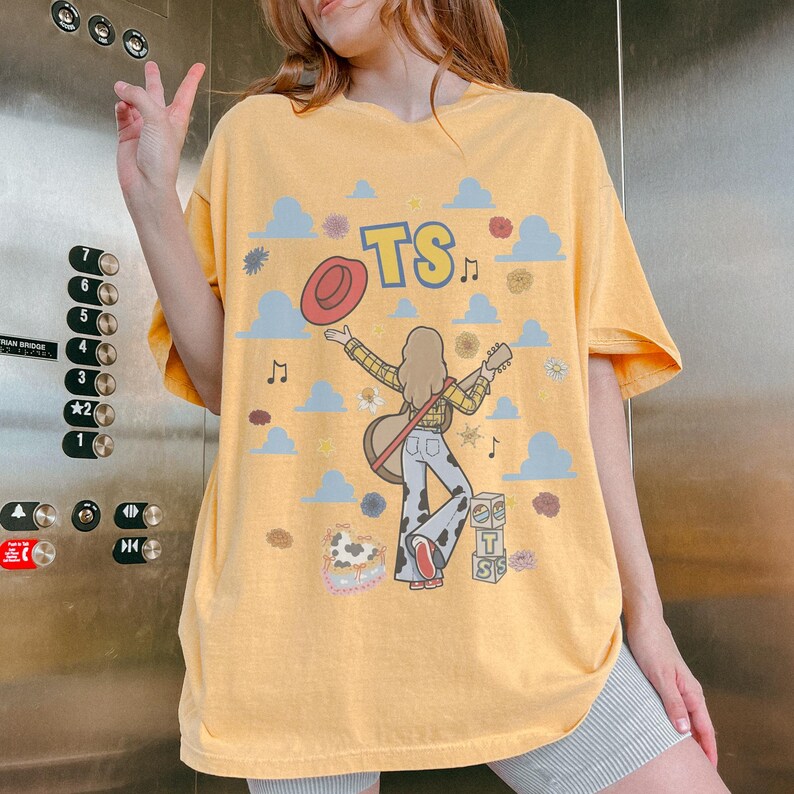 May include: A yellow t-shirt featuring a graphic print of a person with long hair holding a guitar, wearing a yellow plaid shirt and cow-print trousers. The design includes a red hat, blue clouds, musical notes, flowers, and blocks with the text TS.