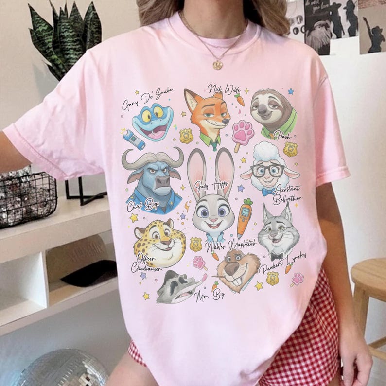 K&ouml;nnte beinhalten: Ein hellrosa T-Shirt mit kurzen &Auml;rmeln und einem Grafikdruck verschiedener animierter Tierfiguren aus Zootopia, darunter Nick Wilde, Judy Hopps und Flash, deren Namen in Schreibschrift neben kleinen Karottensymbolen aufgedruckt sind.