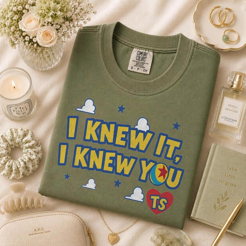 Pu&ograve; includere: Una t-shirt a girocollo color verde oliva con il testo I KNEW IT, I KNEW YOU in lettere gialle e blu, accompagnata da piccole icone di nuvole bianche, stelle blu, una grafica a palla colorata e un cuore rosso contenente le lettere TS.