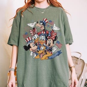 May include: An olive green t-shirt with a graphic of Disney characters Mickey Mouse, Minnie Mouse, Donald Duck, Daisy Duck, Goofy, and Pluto, in patriotic outfits. The design includes American flags and fireworks, with text reading "AMERICA 250th USA Anniversary 1776-2026".