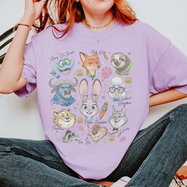 K&ouml;nnte beinhalten: Ein hellviolettes T-Shirt mit einem Grafikdruck verschiedener Zootopia-Figuren, darunter Judy Hopps, Nick Wilde, Flash, Chief Bogo, Assistant Bellwether, Officer Clawhauser, Mr. Big und andere, deren Namen in Schreibschrift aufgedruckt sind.