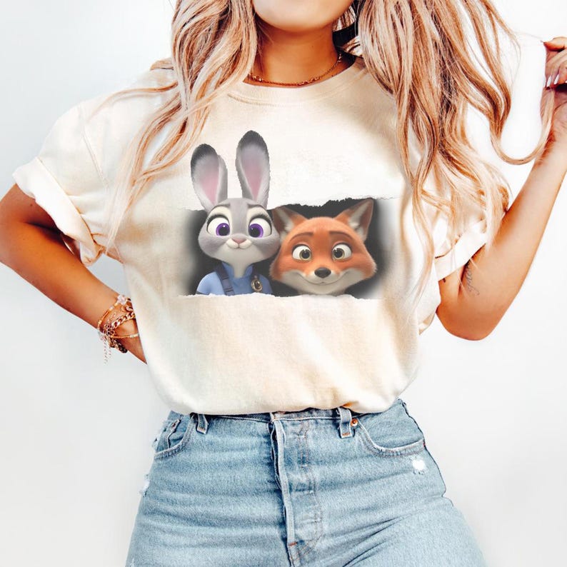 K&ouml;nnte beinhalten: Cremefarbenes T-Shirt mit einer Grafik von Judy Hopps und Nick Wilde aus Zoomania. Die Zeichentrickfiguren sind dargestellt, wie sie aus einem zerrissenen Papiereffekt herausschauen. Das Shirt wird mit blauen Jeans kombiniert.