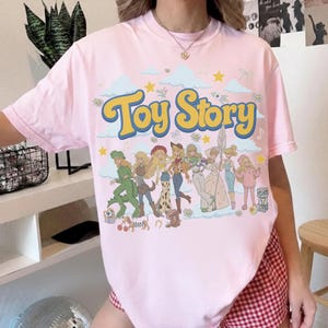 May include: A light pink short-sleeve t-shirt featuring a large graphic print with the text "Toy Story" in yellow letters. The design includes a group of illustrated characters in a retro style, surrounded by blue clouds, yellow stars, and small flowers.