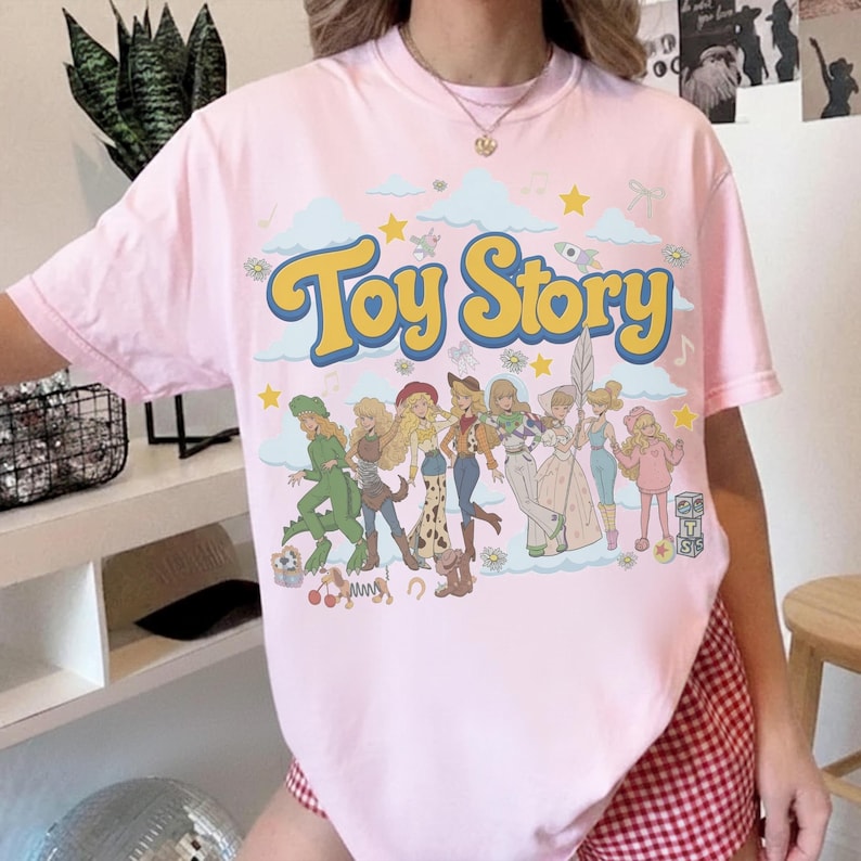 May include: A light pink short-sleeve t-shirt featuring a large graphic print with the text "Toy Story" in yellow letters. The design includes a group of illustrated characters in a retro style, surrounded by blue clouds, yellow stars, and small flowers.