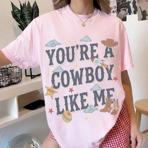 May include: A light pink short-sleeve t-shirt featuring the dark blue text "YOU'RE A COWBOY LIKE ME" surrounded by graphic illustrations of a rocket, a sheriff star, a cowboy hat, cowboy boots, a ball, clouds, and small stars.