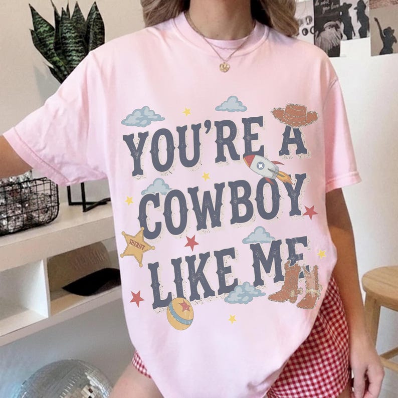 May include: A light pink short-sleeve t-shirt featuring the dark blue text "YOU'RE A COWBOY LIKE ME" surrounded by graphic illustrations of a rocket, a sheriff star, a cowboy hat, cowboy boots, a ball, clouds, and small stars.