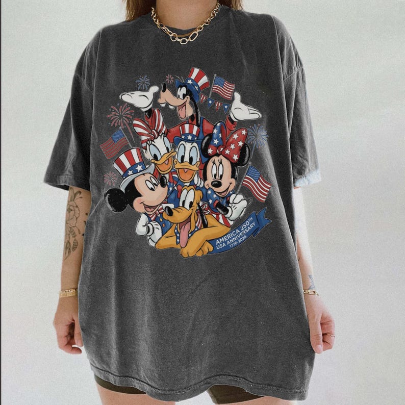 May include: Dark gray t-shirt featuring a colorful graphic of Disney characters Mickey Mouse, Minnie Mouse, Donald Duck, Goofy, and Pluto, with American flags and fireworks. The text "AMERICA 250th USA ANNIVERSARY 1776-2026" is displayed on a blue banner.