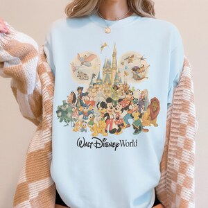 May include: Light blue sweatshirt featuring a colorful graphic of various Disney characters, including Mickey Mouse, Donald Duck, and Simba, in front of a castle. The words "Walt Disney World" are printed in a script font below the characters.