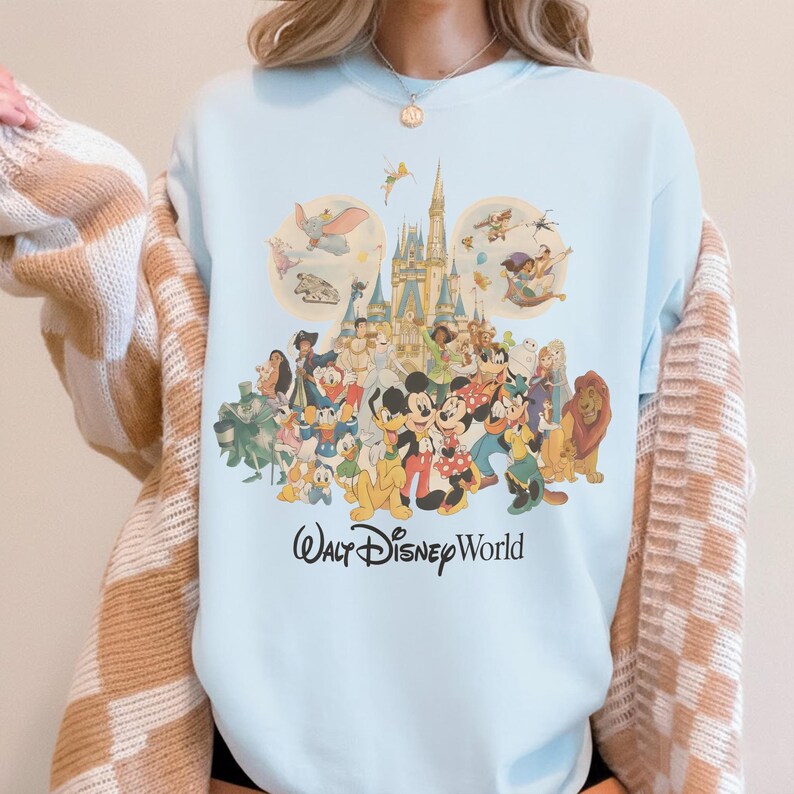 May include: Light blue sweatshirt featuring a colorful graphic of various Disney characters, including Mickey Mouse, Donald Duck, and Simba, in front of a castle. The words "Walt Disney World" are printed in a script font below the characters.