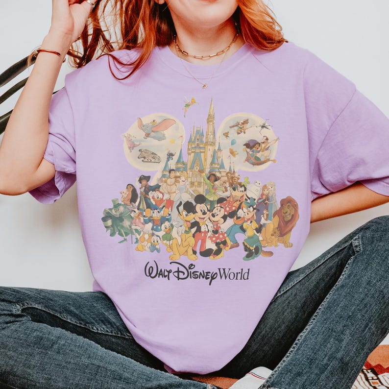 May include: A lilac-colored t-shirt featuring a colorful graphic of Disney characters, including Mickey and Minnie Mouse, Donald Duck, and Simba, in front of a castle. The words "Walt Disney World" are printed below the graphic.