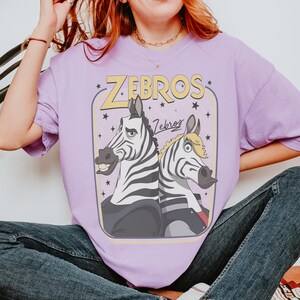 K&ouml;nnte beinhalten: Ein hellviolettes T-Shirt mit einer grafischen Illustration von zwei Zebras, die R&uuml;cken an R&uuml;cken stehen. Der Text "ZEBROS" erscheint in gro&szlig;en gelben Buchstaben oben, mit dem Wort "Zebros" in Schreibschrift darunter, umgeben von kleinen schwarzen Sternen.