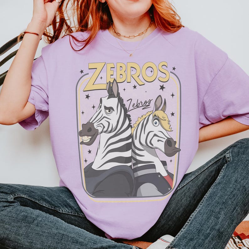 K&ouml;nnte beinhalten: Ein hellviolettes T-Shirt mit einer grafischen Illustration von zwei Zebras, die R&uuml;cken an R&uuml;cken stehen. Der Text "ZEBROS" erscheint in gro&szlig;en gelben Buchstaben oben, mit dem Wort "Zebros" in Schreibschrift darunter, umgeben von kleinen schwarzen Sternen.