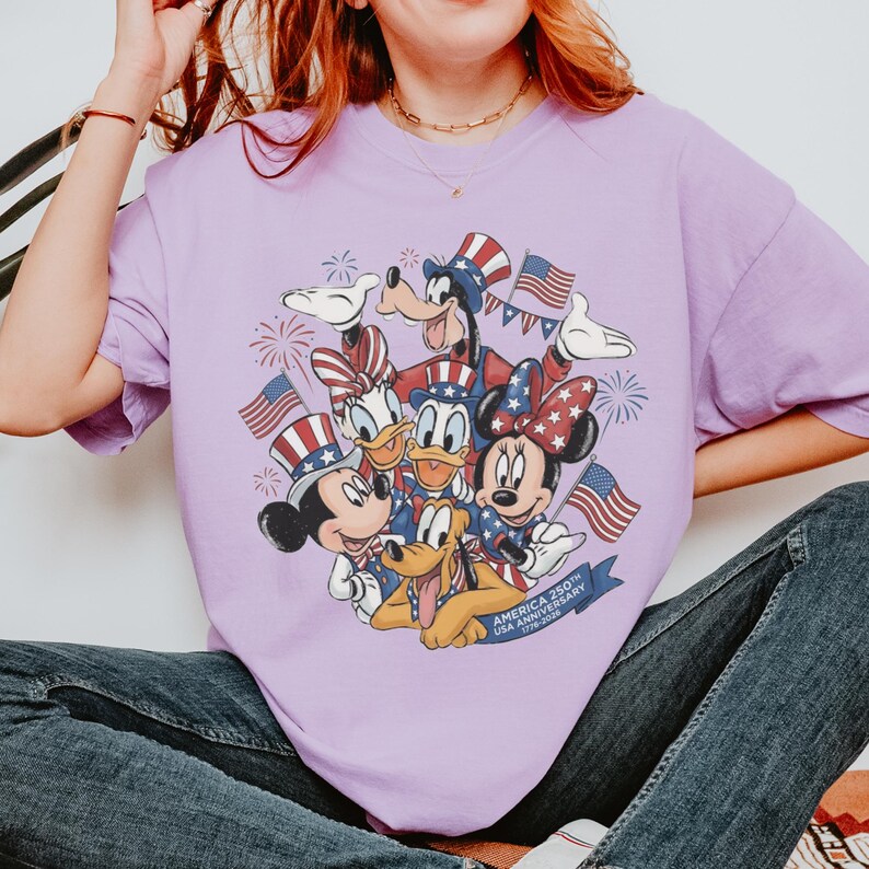 May include: A lilac t-shirt with a graphic of Disney characters Mickey Mouse, Minnie Mouse, Donald Duck, Daisy Duck, Goofy, and Pluto. They are dressed in patriotic outfits, holding American flags, and surrounded by fireworks. The text "AMERICA 250TH USA ANNIVERSARY 1776-2026" is on a blue banner.