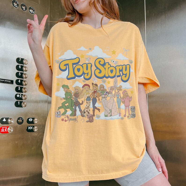 May include: A yellow short-sleeve t-shirt featuring the text "Toy Story" in large blue letters. The graphic includes a group of illustrated characters in various outfits, surrounded by white clouds, small stars, and flowers on a light background.