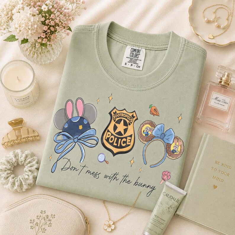 May include: A sage green crewneck t-shirt featuring a graphic print of Zootropolis police elements, including a badge, mouse ears, and a headband with the text "Don't mess with the bunny" in black script. The shirt has a Comfort Colors brand label.