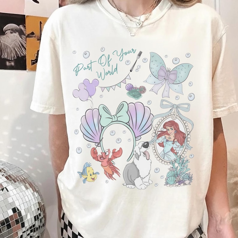 May include: A cream-colored short-sleeve t-shirt featuring a graphic print with the text Part Of Your World, a mermaid, a crab, a fish, a dog, a bow, and seashell-shaped ears. The design includes pastel purple, blue, and green accents.