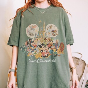 May include: Green t-shirt featuring a colorful graphic of Disney characters and a castle. The design includes Mickey and Minnie Mouse, Donald Duck, and other beloved characters. The words "Walt Disney World" are printed below the graphic.
