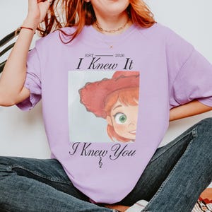 May include: A light purple t-shirt featuring a graphic of a cartoon character with red hair and a red hat. The text reads EST 2026, I Knew It, and I Knew You, with a small musical treble clef symbol integrated into the bottom line of text.