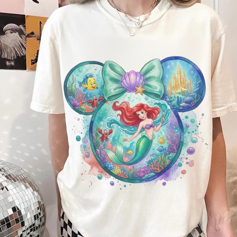 May include: Cream-colored t-shirt featuring a colorful graphic of Ariel the Little Mermaid inside a Mickey Mouse head shape. The design includes Flounder, Sebastian, a castle, and a teal bow with a seashell.